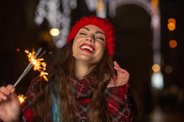 Roześmiana kobieta w czerwonym kapeluszu bawi się wieczorem przy błyszczących światłach na świeżym powietrzu