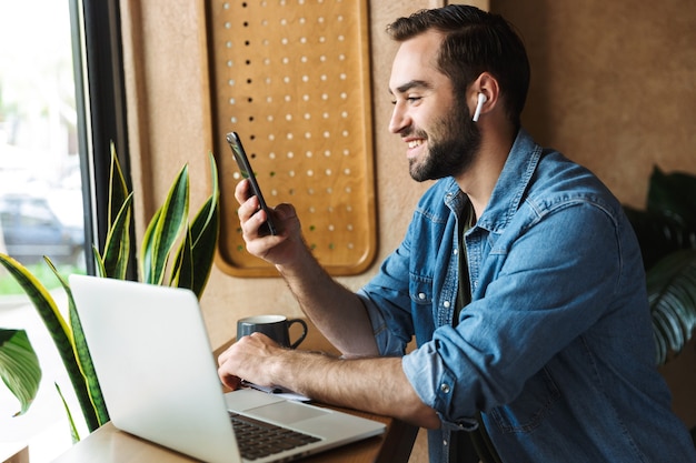 roześmiany kaukaski mężczyzna ubrany w dżinsową koszulę za pomocą słuchawki i telefonu komórkowego z laptopem podczas pracy w kawiarni w pomieszczeniu