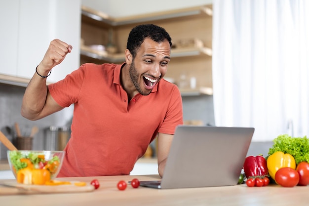 Roześmiany podekscytowany Afroamerykanin z Tysiąclecia przy stole z ekologicznymi warzywami używa laptopa i wykonuje gest sukcesu