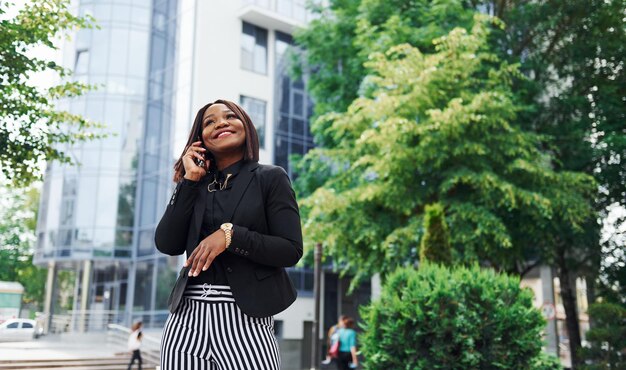 Rozmawia przez telefon Młoda afroamerykańska kobieta w modnych ubraniach na zewnątrz w mieście w pobliżu zielonych drzew i przeciwko budynkowi biznesowemu