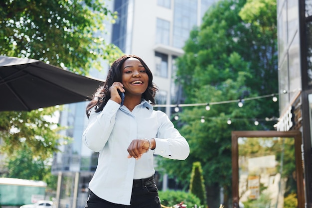Rozmowa za pomocą telefonu Młoda afro amerykańska kobieta w białej koszuli na zewnątrz w mieście w pobliżu zielonych drzew i przeciwko budynkowi biznesowemu