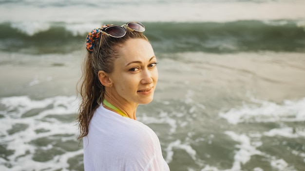 Rozmycie mgły blask filmowy zbliżenie portret kobiety w białej koszuli tuniki na plaży w pobliżu wzburzonego morza Atrakcyjna dziewczyna z mokrymi kręconymi włosami w okularach przeciwsłonecznych Kobiece spacery turystyczne bawią się odpoczynek o zachodzie słońca