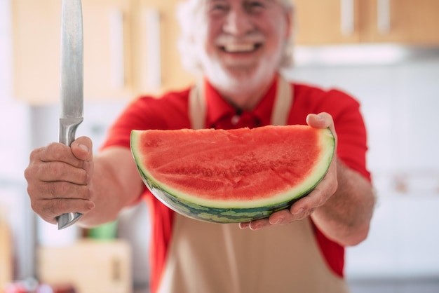 Rozmyty starszy mężczyzna w kuchni trzymający nóż i arbuza aktywni uśmiechnięci emeryci cieszący się zdrowym i wegetariańskim jedzeniem