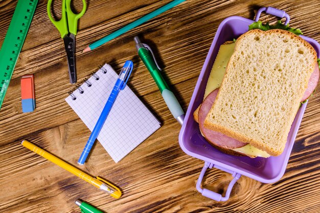 Różne artykuły papiernicze i pudełko na lunch z kanapkami na drewnianym stole Widok z góry