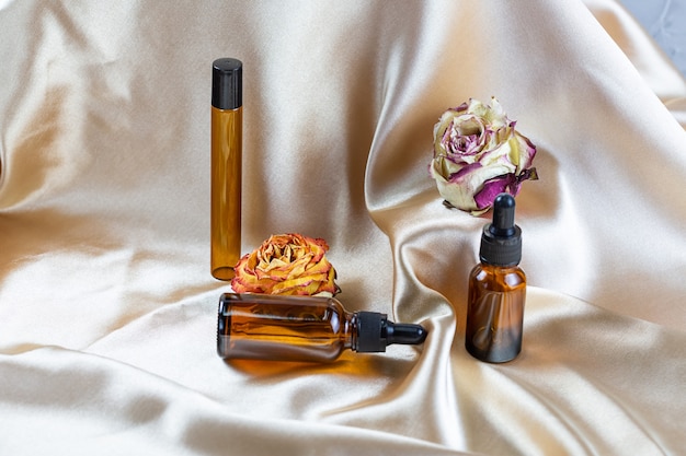 Różne pojemniki do przechowywania kosmetyków z ciemnego szkła leżą na fałdach jedwabnej satynowej tkaniny, otoczone suszonymi kwiatami róży. Butelka perfum, serum lub balsamu