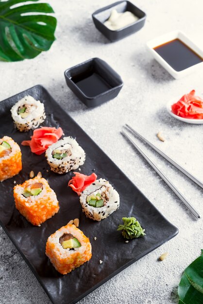 Różne Świeże i pyszne sushi ustawione na czarno-białym łupku z metalowymi i łupkowymi pałeczkami, sosem i zielonymi liśćmi na jasnej kamiennej powierzchni