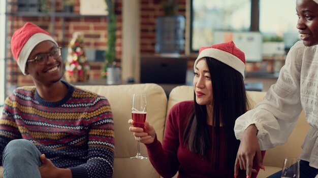 Różnorodni pracownicy piją kieliszki wina na przyjęciu bożonarodzeniowym w biurze ozdobionym świątecznymi ozdobami. Serwowanie napojów alkoholowych podczas ferii zimowych w czapkach Mikołaja. Strzał z ręki.