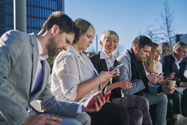 różnorodni współpracownicy podczas przerwy poza biurem siedzą na ławce i bawią się za pomocą smartfona