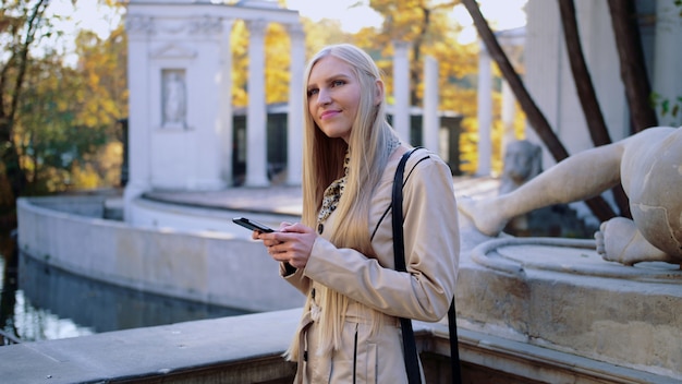 Rozochocona blondynki kobieta w beżowym żakiecie w jesień parku, patrzeje jej smartphone