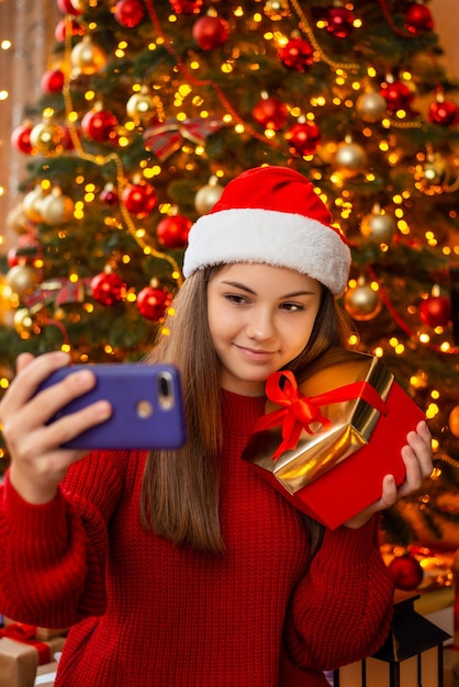 Rozochocona młoda dziewczyna w santa hat robi selfie z prezenta pudełkiem blisko choinki