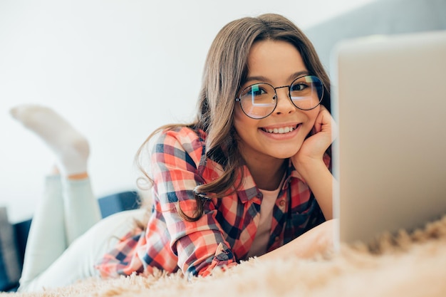 Rozochocona nastolatka w okularach leżąca na łóżku przed laptopem z dłonią dotykającą policzka