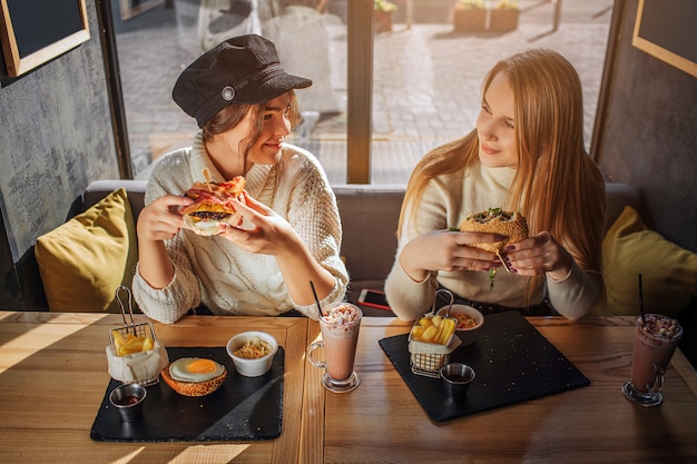 Zdjęcie rozochocone młode kobiety patrzeją eahc inny i one uśmiechają się. siedzą przy stoliku w kawiarni i trzymają hamburgery. młode kobiety są szczęśliwe.