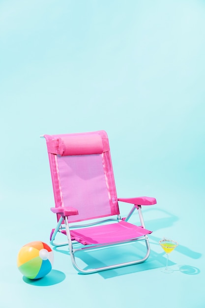Różowe krzesło plażowe, kolorowa piłka plażowa i żółty napój w eleganckim szkle na jasnoniebieskim tle