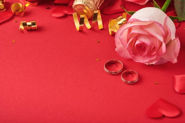 Różowe kwiaty róż, obrączki, szampan, prezent, złote wstążki i konfetti serca na czerwonym tle. Widok z góry z miejscem na pozdrowienia. Walentynki lub wesele tło i kartkę z życzeniami.
