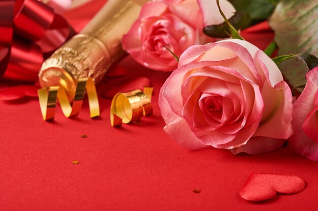 Różowe kwiaty róż, szampan, prezent, złote wstążki i konfetti czerwone serca na czerwonym tle. Płaski widok z góry z miejscem na pozdrowienia. Walentynki tło i kartkę z życzeniami.