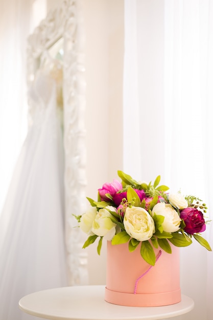 Różowe okrągłe pudełko z kwiatami, wewnątrz kolorowych piwonii na tle białego lustra z suknią ślubną