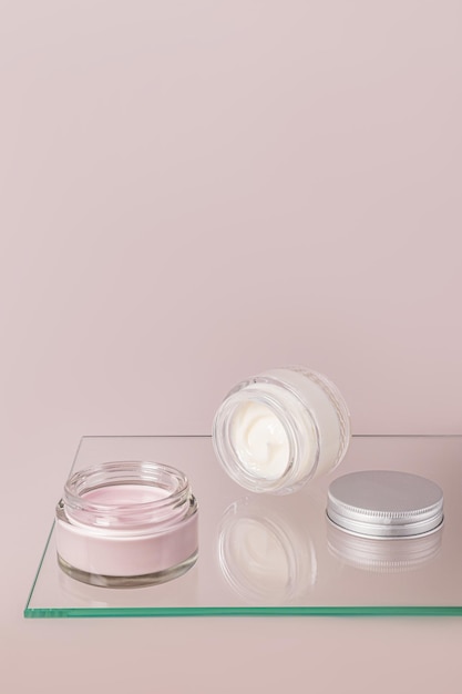 Zdjęcie różowy i biały krem do pielęgnacji skóry twarzy i ciała w otwartych szklanych słoikach kosmetycznych na szklanej powierzchni pływająca półka widok pionowy miejsce do kopiowania