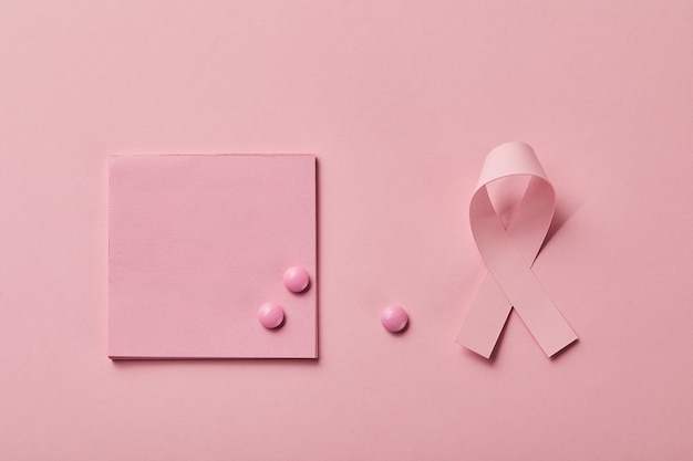 Rozproszone pigułki na kartce papieru w pobliżu wstążki raka piersi na jasnoróżowym tle