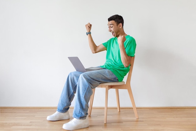 Rozradowany Arab używający laptopa, potrząsając pięściami i krzycząc, siedząc na krześle nad wolną przestrzenią na jasnej ścianie