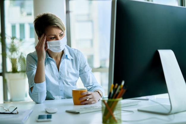 Rozważna bizneswoman nosząca maskę podczas picia kawy w biurze