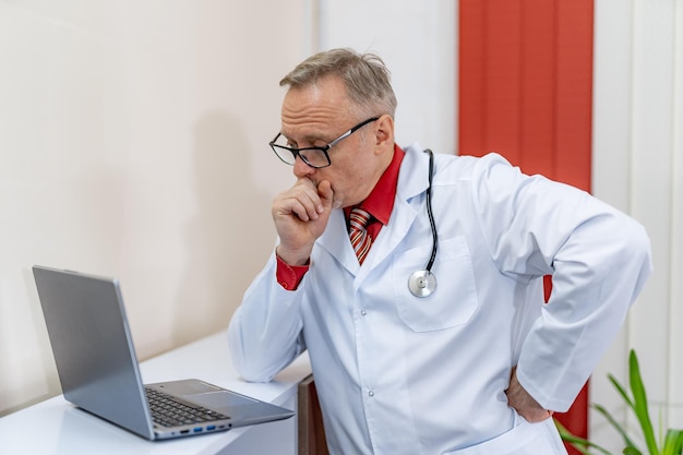 Zdjęcie rozważny dojrzały lekarz z laptopem na biurku w klinice. medyk myśli o przyszłym wyleczeniu pacjenta.