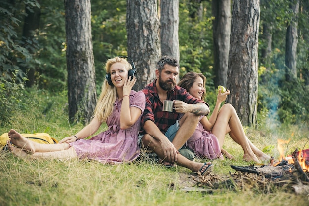 Rozważny mężczyzna z brodą pije herbatę z kawą, obserwując płomienie ogniska. Blond dziewczyna słucha muzyki, podczas gdy jej siostra je jabłko. Różne postrzeganie idealnego relaksu.