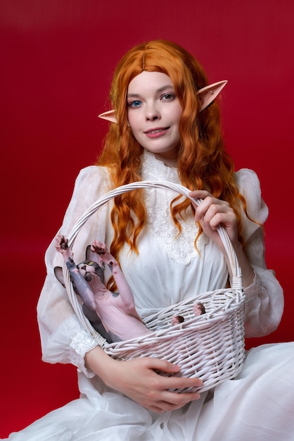 Ruda cosplayerka elf młoda kobieta w białej sukni trzyma biały wiklinowy kosz z zabawnym kotem sfinksem c