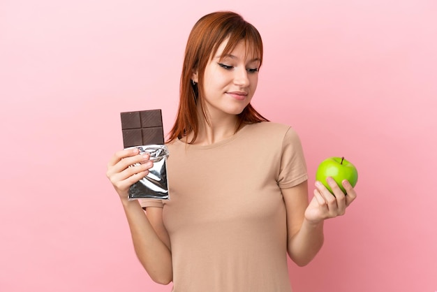 Ruda dziewczyna odizolowana na różowym tle bierze w jedną rękę tabliczkę czekolady, a w drugiej jabłko