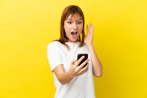 Ruda dziewczyna odizolowana na żółtym tle patrząca w kamerę podczas korzystania z telefonu komórkowego ze zdziwionym wyrazem twarzy