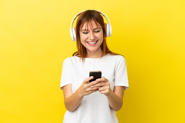 Ruda dziewczyna odizolowana na żółtym tle słuchająca muzyki i patrząca na telefon komórkowy