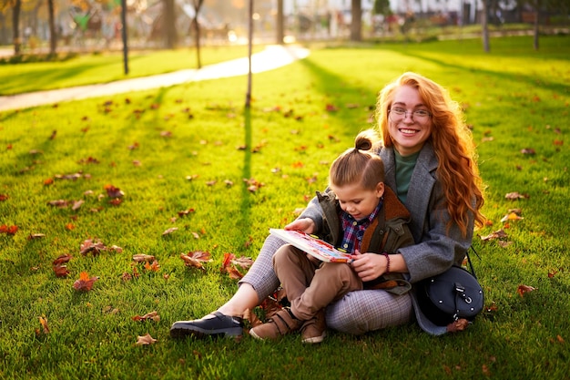 Ruda kobieta czyta książkę z małym chłopcem siedzącym na trawie w parku miejskim Młoda mama uczy swojego pierwszego równiarki syna i odrabia pracę domową w słoneczny jesienny dzień Powrót do koncepcji szkoły Liście na zielonym trawniku