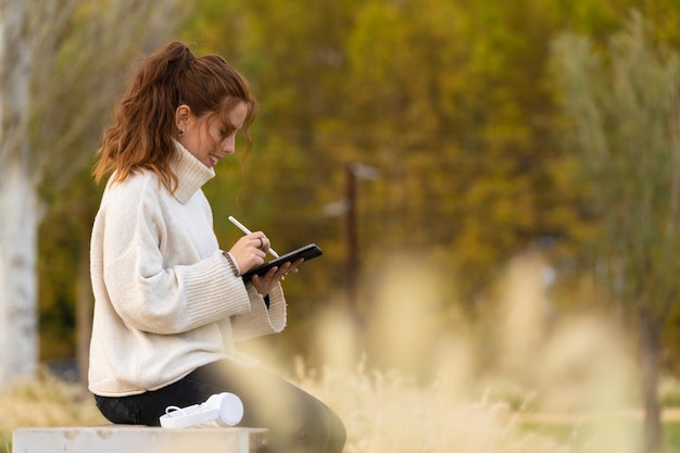 Ruda młoda kobieta pisze elektronicznym piórem na tablecie i w parku Ma na sobie biały sweter z golfem