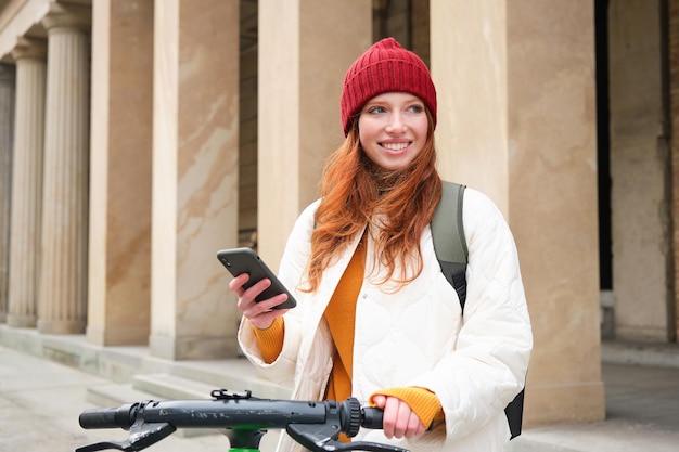 Ruda turystka z plecakiem używa telefonu komórkowego do wynajęcia skutera na ulicach europejskiego miasta