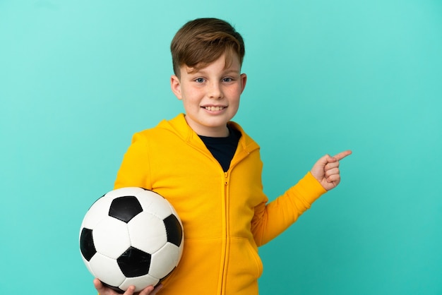 Rude dziecko gra w piłkę nożną na białym tle na niebieskiej ścianie, wskazując palcem w bok