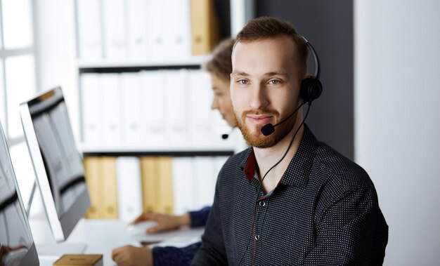 Rudobrody biznesmen rozmawia przez zestaw słuchawkowy w pobliżu swojej koleżanki, siedząc w nowoczesnym biurze. Grupa różnych ludzi w call center. Telemarketing i obsługa klienta.