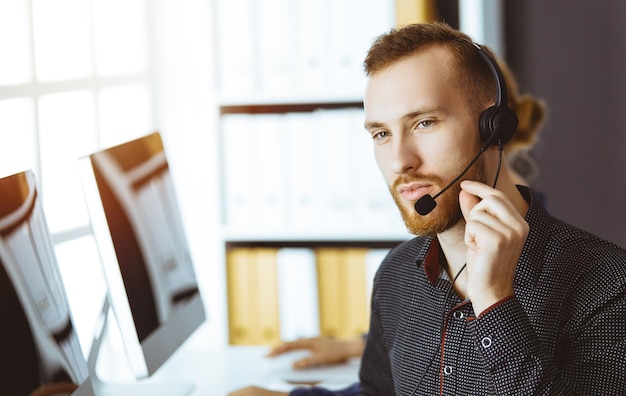 Rudobrody biznesmen rozmawia przez zestaw słuchawkowy w pobliżu swojej koleżanki, siedząc w nowoczesnym, słonecznym biurze Zróżnicowana grupa ludzi w call center