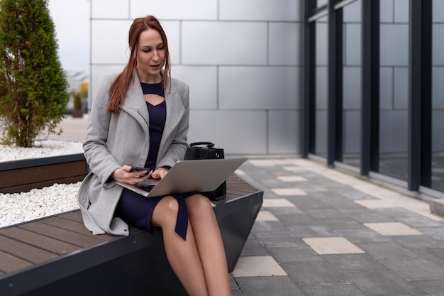 Rudowłosa bizneswoman w ciuchach biznesowych pracuje na laptopie online w pobliżu biura biznesowego