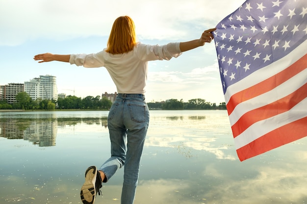 Zdjęcie rudowłosa dziewczyna trzyma flagę narodową usa w jej ręce. pozytywna młoda kobieta z okazji dnia niepodległości stanów zjednoczonych.