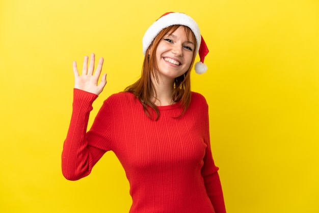 Rudowłosa dziewczyna w bożonarodzeniowym kapeluszu na żółtym tle pozdrawiająca ręką ze szczęśliwym wyrazem twarzy