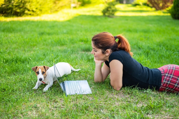 Rudowłosa kobieta leży na zielonej trawie w parku ze szczeniakiem Dziewczyna zachowuje dystans społeczny i ogląda wideo na świeżym powietrzu na laptopie Student uczy się zdalnie na komputerze