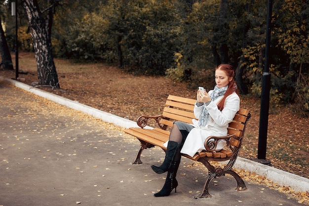 Rudowłosa kobieta w średnim wieku ze smartfonem w ręku siedzi na ławce w parku