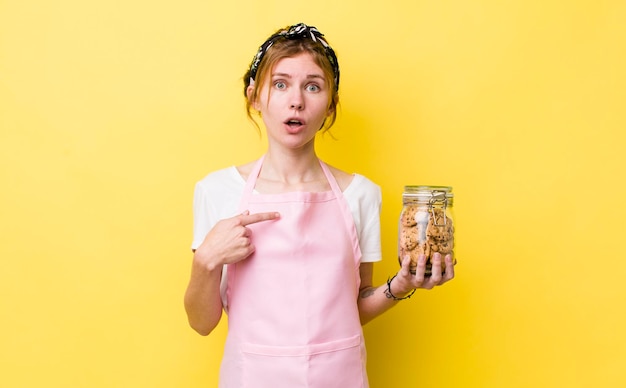 Zdjęcie rudowłosa ładna kobieta wygląda na zszokowaną i zaskoczoną z szeroko otwartymi ustami, wskazując na gospodynię domową trzymającą butelkę ciastek