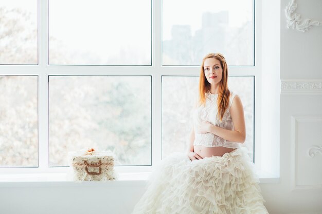 Rudowłosa młoda dziewczyna w ciąży w białej sukience przy oknie
