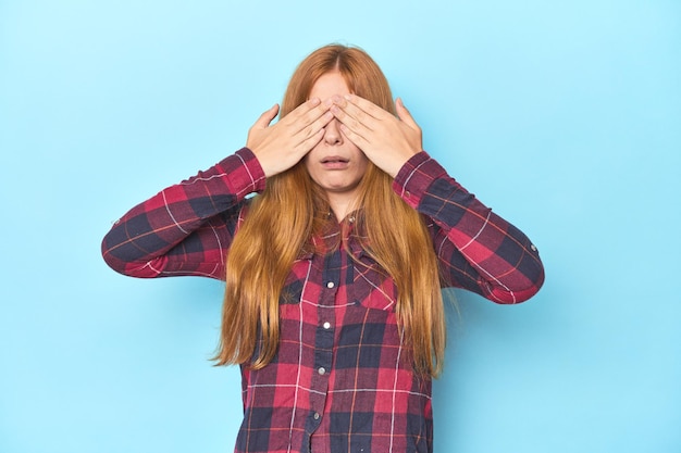 Zdjęcie rudowłosa młoda kobieta na niebieskim tle boi się zakrywać oczy rękami