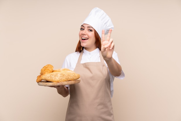 Rudowłosa nastolatka dziewczyna w mundurze szefa kuchni. Żeński piekarz trzyma stół z kilka chlebami uśmiecha się zwycięstwo znaka i pokazuje