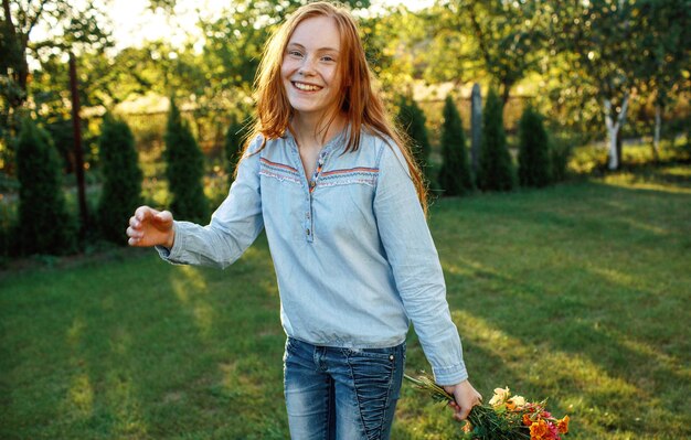 Rudowłosa Nastolatka Tańczy Na Trawniku Z Bukietem Kwiatów W Dłoni. Słoneczny Zachód Słońca