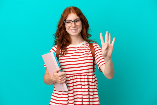 Rudowłosa studentka nastolatka na białym tle na niebieskim tle szczęśliwa i licząca cztery palcami