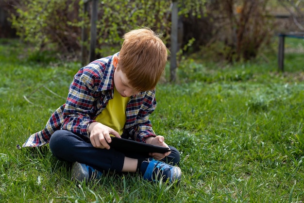 Rudy chłopiec bawi się na tablecie lub ogląda bajki siedząc na zielonej trawie na podwórku w wiosce