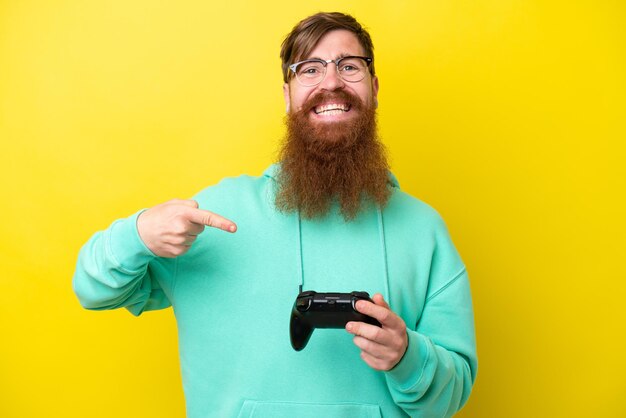 Rudy mężczyzna z brodą bawi się kontrolerem gier wideo na żółtym tle i wskazuje go