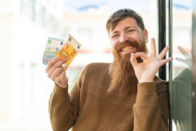 Zdjęcie rudy mężczyzna z brodą, biorąc dużo pieniędzy na zewnątrz, pokazując znak ok palcami
